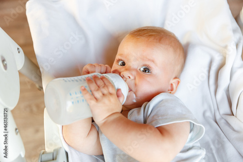Cute little newborn girl drinking milk from bottle and looking at camera on white background. Infant baby sucking eating milk nutrition lying down on crib bed at home. Motherhood happy child concept.
