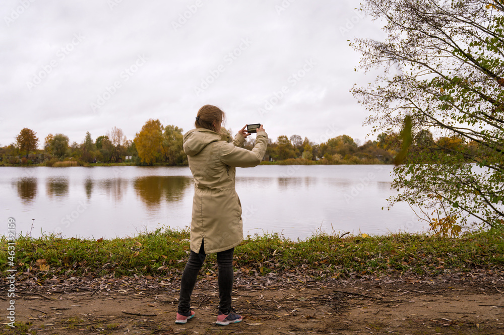 Girl on the shore of the river is taking a picture of autumn nature.