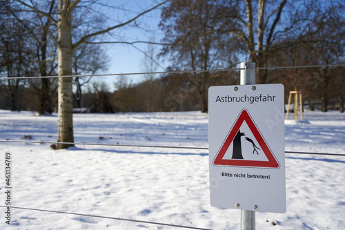 Rectangular white sign on a fence saying: Risk of branch breakage, entry prohibited. (German: Astbruchgefahr, betreten verboten). Bare trees on frozen snow ground. Winter in Germany.