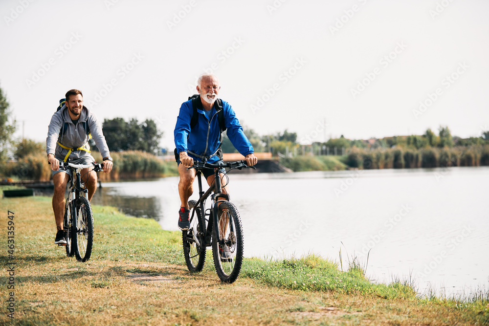 Happy senior athlete and his son ride bicycles by the lake in nature. Copy space. - obrazy, fototapety, plakaty 