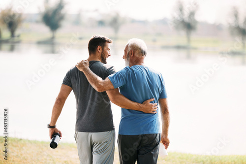 Rear view of athletic father and son talk while walking embraced by the lake.