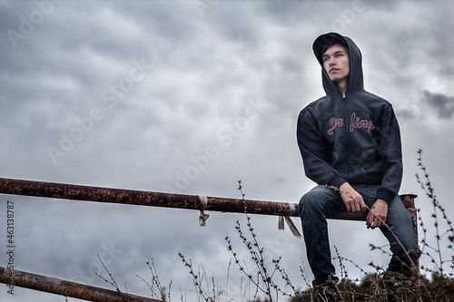 A young guy in a hood walks on a barrier against an overcast gray sky