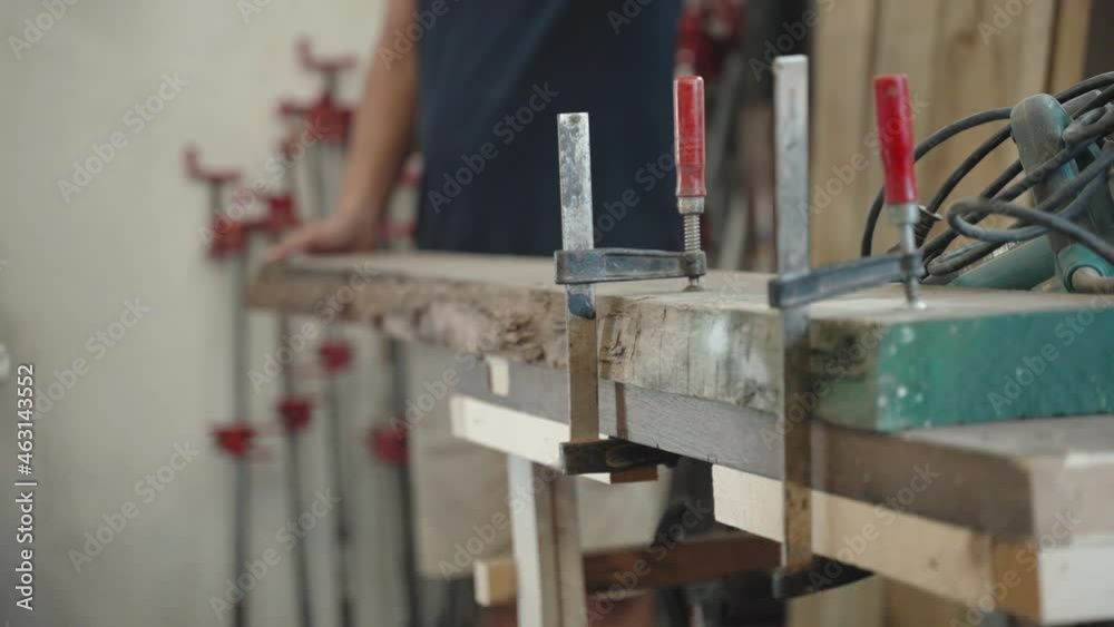 Professional craftsman tightening raw wooden plank with steel clamps to the workbench for woodworking process, handyman using electric grinding machine for smoothing wooden surface texture before