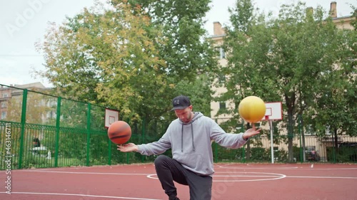 Skilled young man doing basketball freestyle trick spinning two balls on index fingers and kneeling on outdoor college court. Young male freestyler performing stunt with two basketballs photo