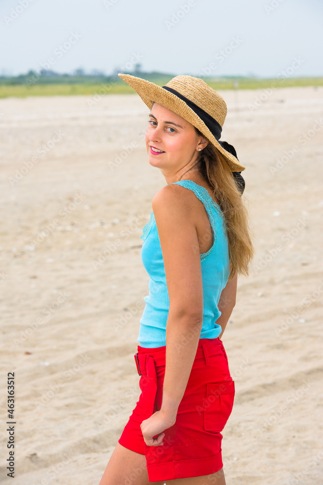 A pretty blonde woman, dressing in light blue tank top and red shorts, wearing a yellow straw hat, is charmingly standing on a sandy beach.. A pretty blonde woman, dressing in light blue tank top and red shorts, wearing a yellow straw hat, is charmingly standing on a sandy beach..