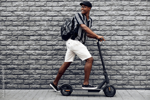 African american man with scooter on black brick wall background, guy with backpack