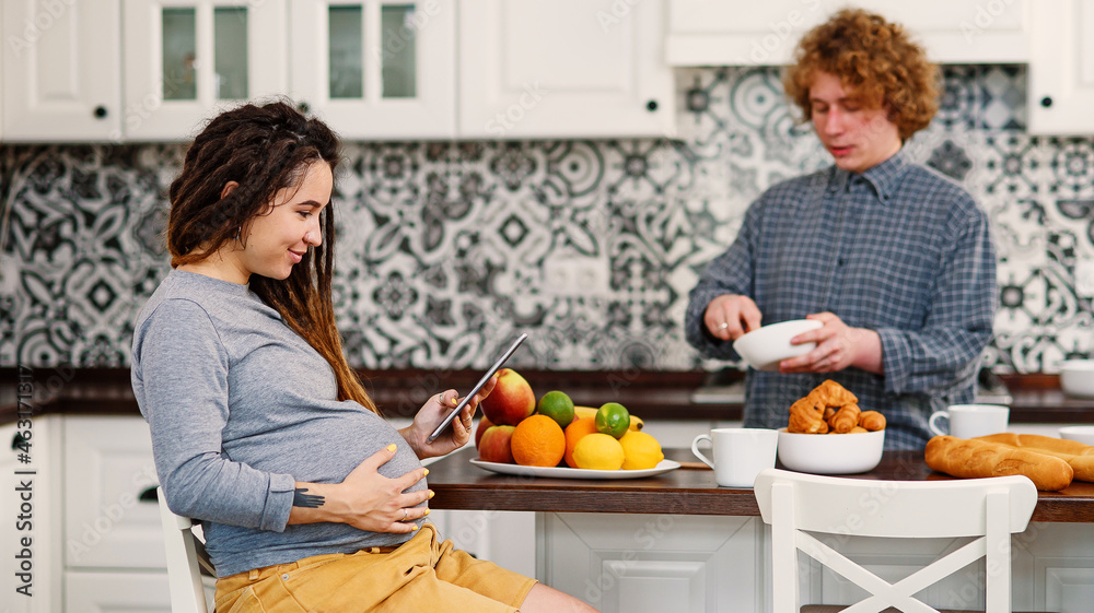 Joyful pregnant woman with dreadlocks drinks tea and watching her curly husband cooking breakfast for her in kitchen