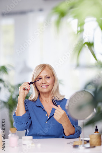 A happy senior woman sitting at home and putting mascara on eyelashes. Senior woman using make-up