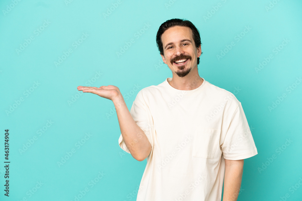 Young caucasian man isolated on blue background holding copyspace imaginary on the palm to insert an ad