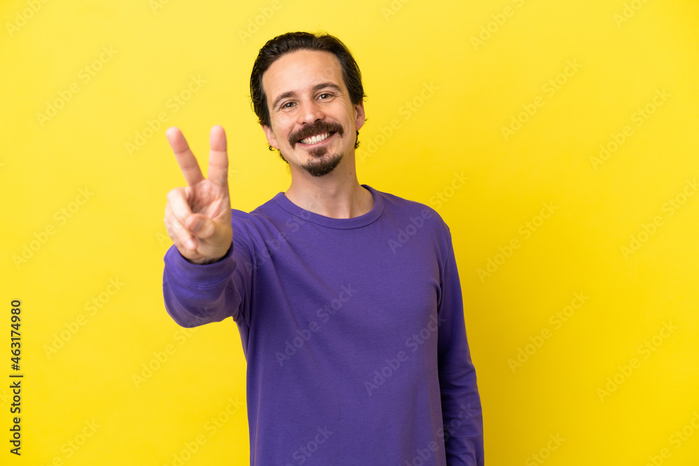 Young caucasian man isolated on yellow background smiling and showing victory sign