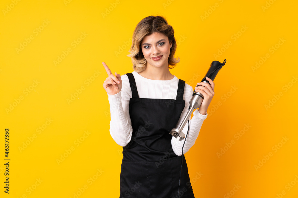 Girl using hand blender isolated on yellow background showing and lifting a finger in sign of the best Girl using hand blender isolated on yellow background showing and lifting a finger in sign of the best