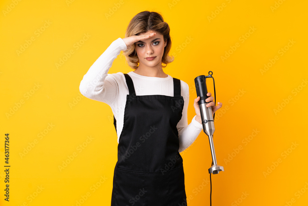 Girl using hand blender isolated on yellow background saluting with hand with happy expression