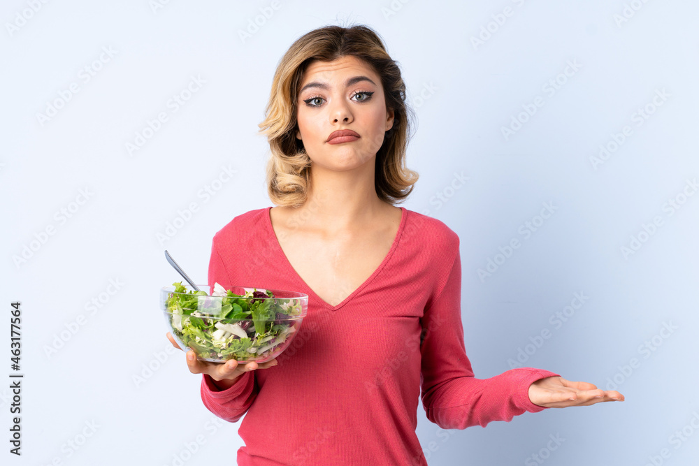Teenager woman with salad isolated on blue background making doubts gesture while lifting the shoulders Teenager woman with salad isolated on blue background making doubts gesture while lifting the shoulders