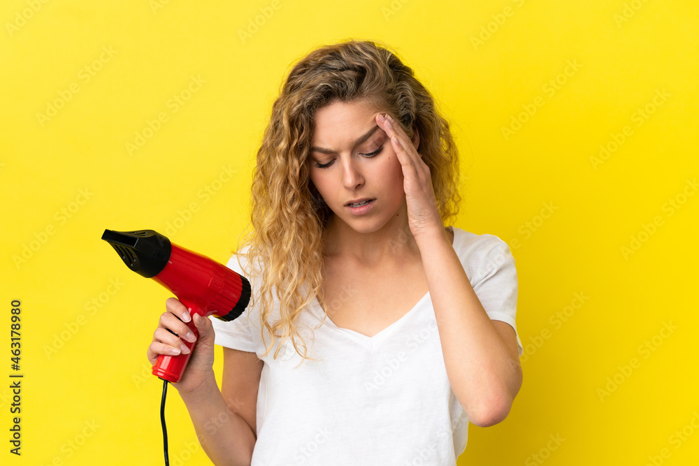 Young blonde woman holding a hairdryer isolated on yellow background with headache Young blonde woman holding a hairdryer isolated on yellow background with headache