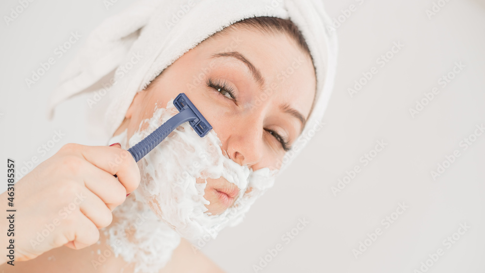 Cheerful caucasian woman with a towel on her head and shaving foam on her face holds a razor on a white background