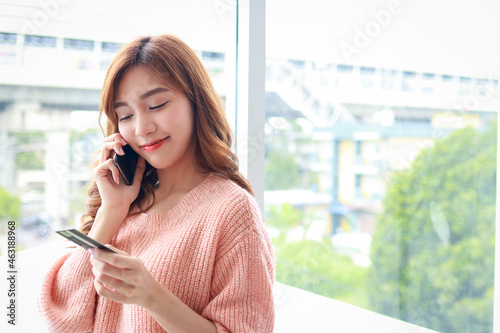 beautiful asian woman Lives in an apartment, standing at a glass window. holding a black smartphone and credit card to order online. Concept of using modern technology in daily life. Work from home.