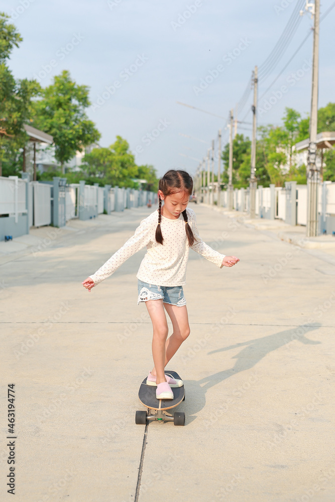 Asian little girl child skating on a skateboard. Kid riding on skateboard outdoors at the street. She skateboarding on the road Asian little girl child skating on a skateboard. Kid riding on skateboard outdoors at the street. She skateboarding on the road