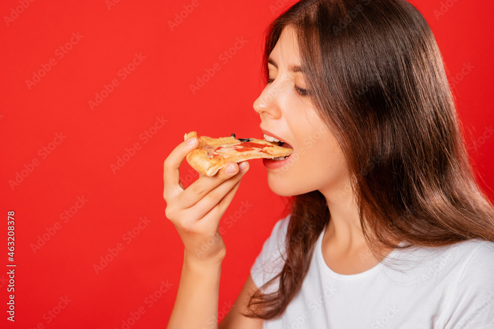 Close up portrait of a young woman in a white t-shirt eating a slice of pizza isolated on a red background. Copy space for label, mockup.