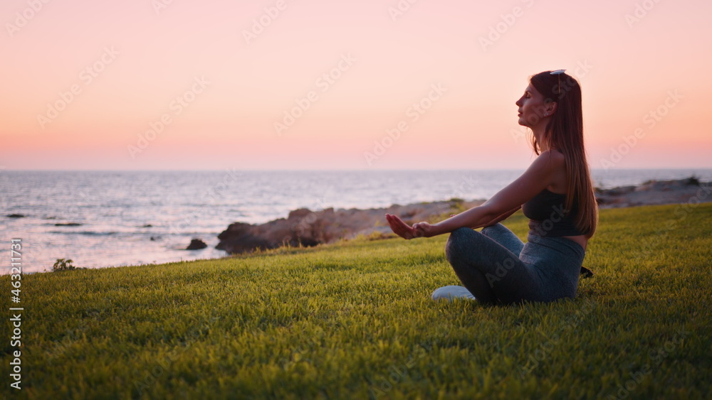 Side view of peaceful caucasian woman in activewear sitting on lotus position on beach grass and meditating with closed eyes. Yoga practises near ocean during evening time. Side view of peaceful caucasian woman in activewear sitting on lotus position on beach grass and meditating with closed eyes. Yoga practises near ocean during evening time.