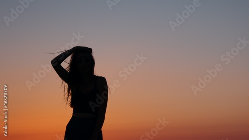 Silhouette of female athlete with slender body resting after evening exercises on beach. Amazing summer sunset over ocean. Enjoyment from outdoors workout.