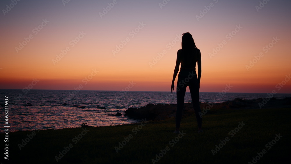 Young woman with slender body practising in fitness exercises during evening time near ocean. Silhouette of active healthy female. Regular training for body care. Woman fitness near ocean.