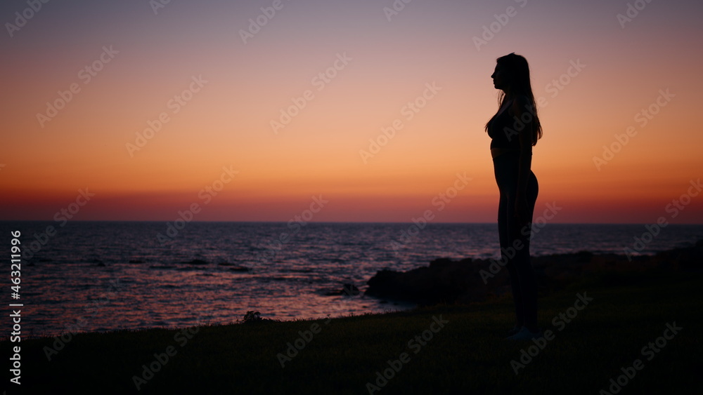 Fitness woman in silhouette doing squats exercises on beach near ocean. Young female in sportswear spending evening time for outdoors activity. Amazing colorful sunset on background.