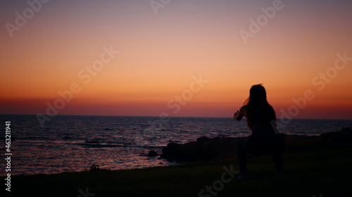 Fitness woman in silhouette doing squats exercises on beach near ocean. Young female in sportswear spending evening time for outdoors activity. Amazing colorful sunset on background. Fitness woman. © GRAFStock