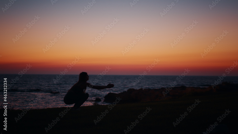 Fitness woman in silhouette doing squats exercises on beach near ocean. Young female in sportswear spending evening time for outdoors activity. Amazing colorful sunset on background. Fitness woman.