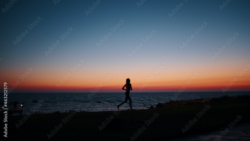 Silhouette of sporty young woman in activewear running along beach during evening time. Physical exercises during summer sunset over ocean. Motivation and endurance concept.