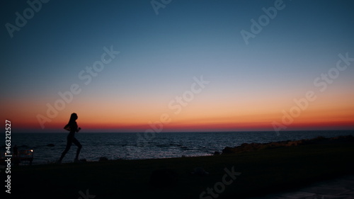 Silhouette of sporty young woman in activewear running along beach during evening time. Physical exercises during summer sunset over ocean. Motivation and endurance concept.