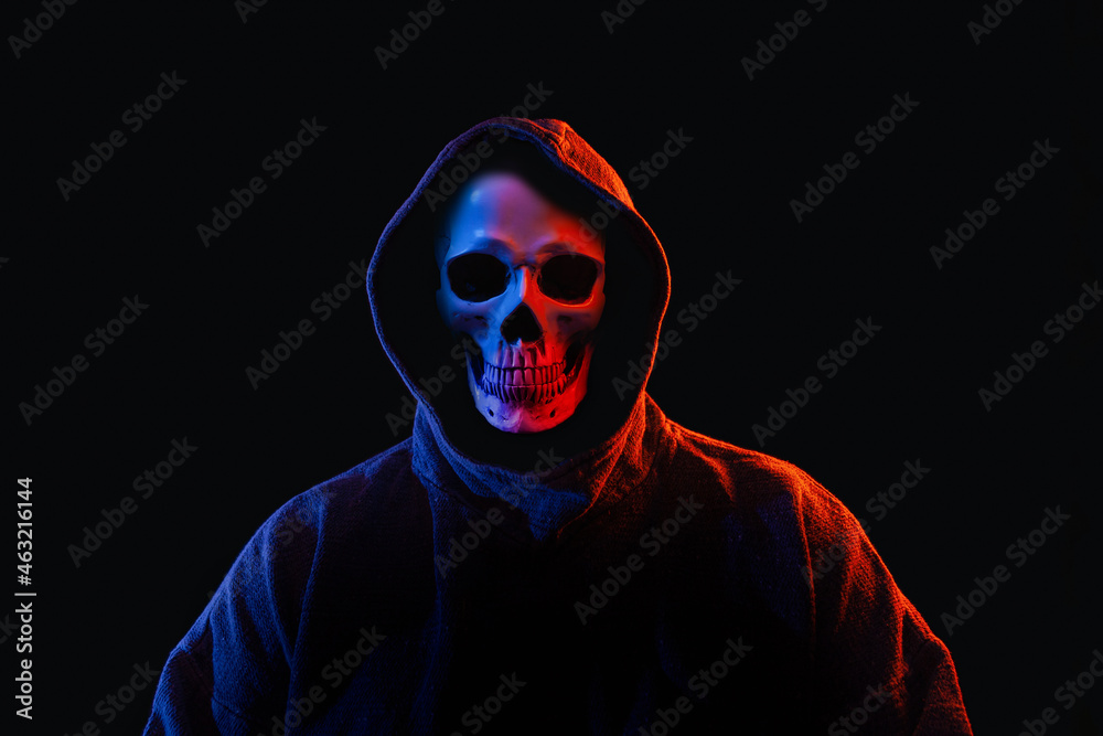 Man with human skull instead of face in dark hoodie illuminated with red and blue lights