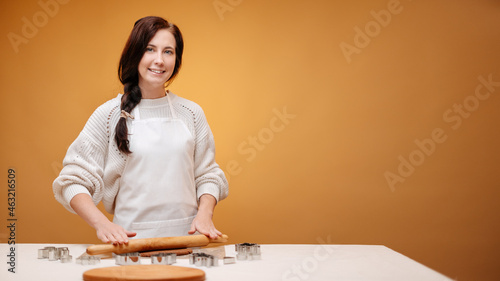 Happy woman baker in a white apron prepares New Year's cookies on a yellow background.