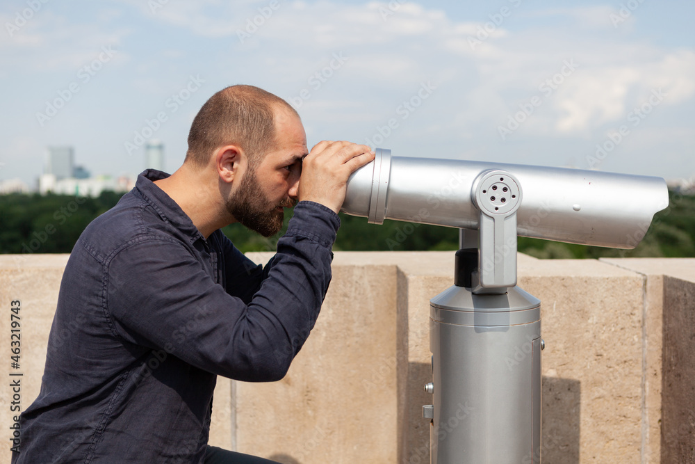 Man tourist standing on building rooftop looking through binocular telescope at metropolitan city admiring beautiful place. Panoramic landscape view from tower observation point.