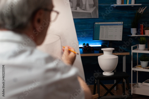 Caucasian artist looking at vase on desk and drawing on canvas at workplace. Senior man with artistic hobby recreating professional masterpiece using art intruments and tools. photo