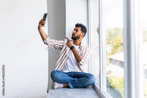 Young indian man sitting on the windowsill make video call on the phone