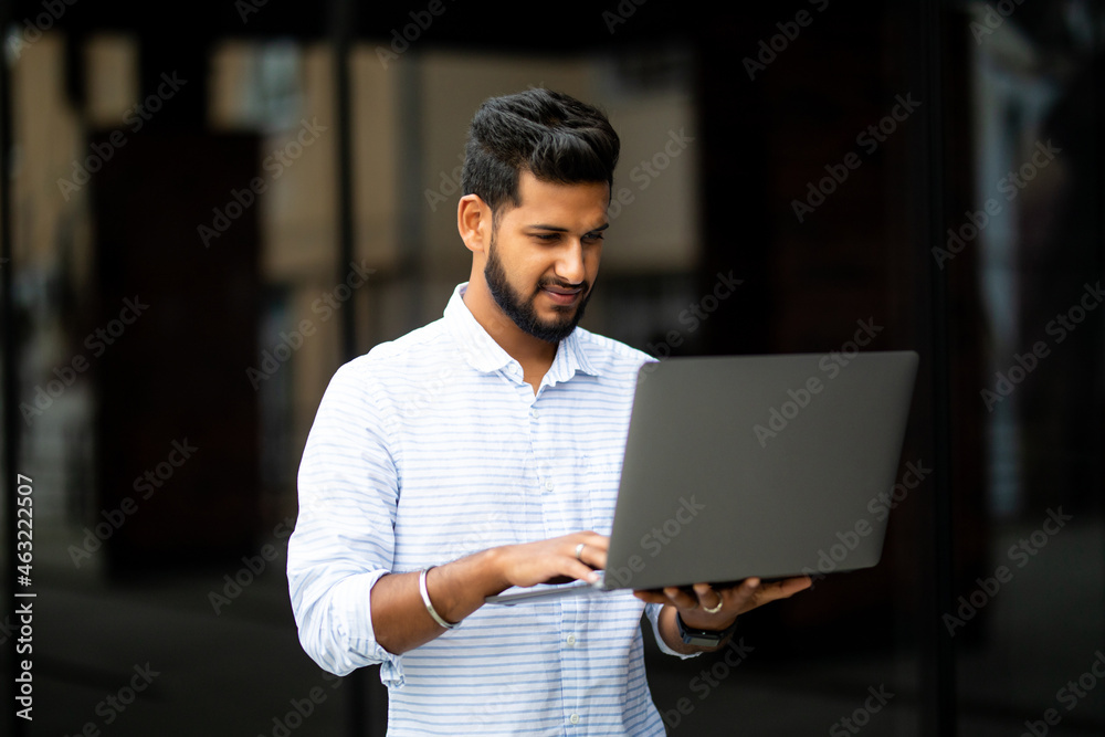 Young indian man working on the computer and drinking coffee on the street opposite the office