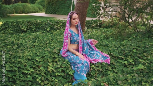 Eastern fantasy woman sits on green grass ivy, vacation meditates, enjoy nature, silence summer tropical garden park. National India blue pink dress saree, scarf on head. Silver teak tiara. Indin girl photo