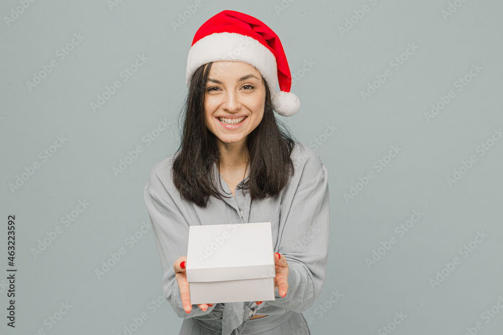 Photo of cute smiling female in Christmas hat is giving present. Wears grey shirt, isolated grey color background Photo of cute smiling female in Christmas hat is giving present. Wears grey shirt, isolated grey color background