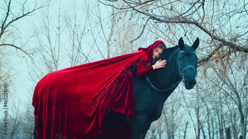 Medieval fantasy queen, horsewoman woman sittin, lies astride black horse hand strokes mare's back. Red long cloak cape. Hood on head redhaired girl princess. Bare tree branches nature winter forest. photo