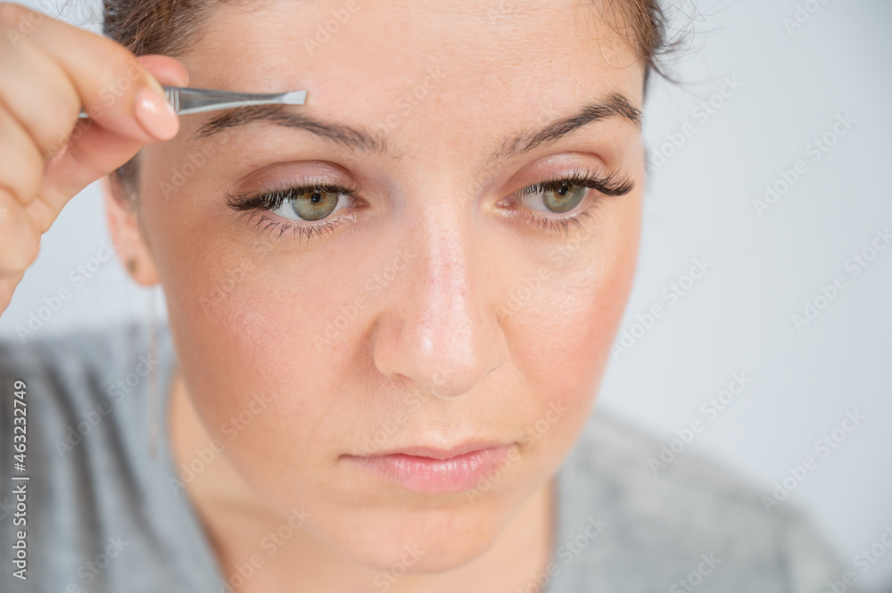 Close-up portrait of a caucasian woman doing eyebrow correction herself with tweezers