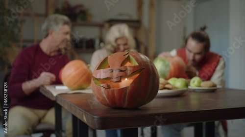 A pumpkin lantern with a candle in its mouth stands at the edge of the table. People in the background are busy preparing for Halloween