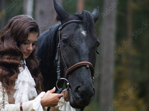 A young beautiful brunette rider next to a black mare in full ammunition,