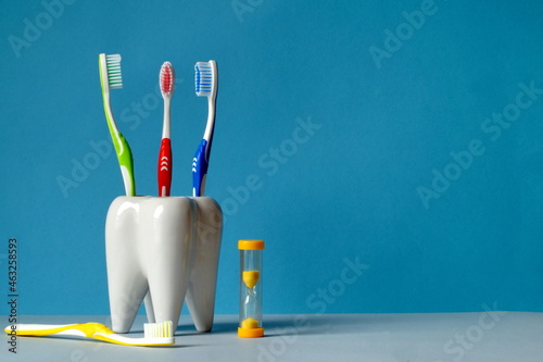 Multi-colored toothbrushes are in a glass, in the shape of a tooth, on the table there is a brush of a very color and yellow hourglass to count the time of brushing your teeth