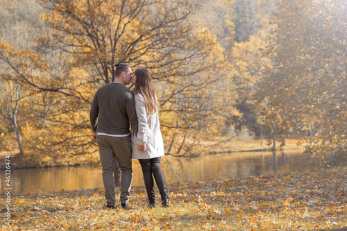 Love, leisure, nature, relationship concept. Young couple walking on meadow with yellow leaves in the park with pond and trees during autumn sunset golden ours. Bright vivid backlight filter
