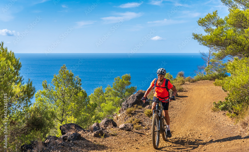 Fototapeta premium nice woman riding her electric mountain bike at the coastline of mediterranean sea on the Island of Elba in the tuscan Archipelago, in front of Porto Ferraio,Tuscany, Italy