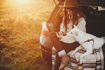Travel and autumn road trip with pet. Stylish hipster woman in sweater holding phone and sitting with cute dog in car trunk in sunset light. Young female with smartphone traveling. Space for text