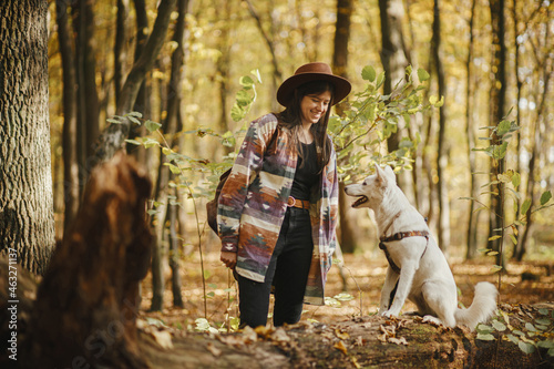 Stylish woman in hat training cute dog sitting on old stump in sunny autumn woods. Young female traveler with swiss shepherd white dog in beautiful forest. Travel and learning with pet