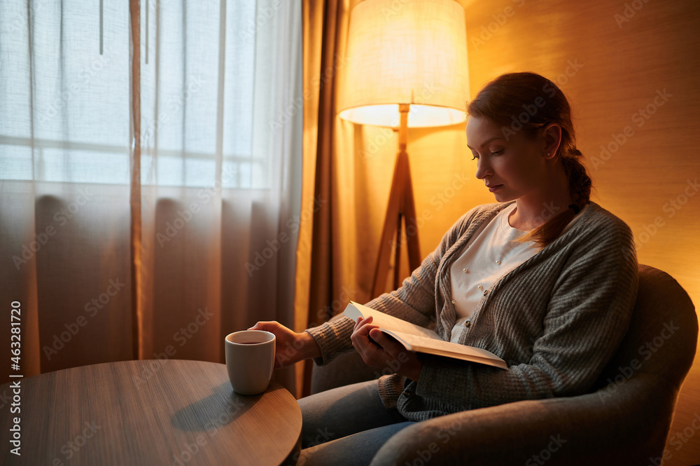 Cozy home environment. Soft yellow light from a floor lamp. Young woman in a chair near the window reads a book. She has a cup of coffee in her hand. Hotel, home interior. Cozy home environment. Soft yellow light from a floor lamp. Young woman in a chair near the window reads a book. She has a cup of coffee in her hand. Hotel, home interior.