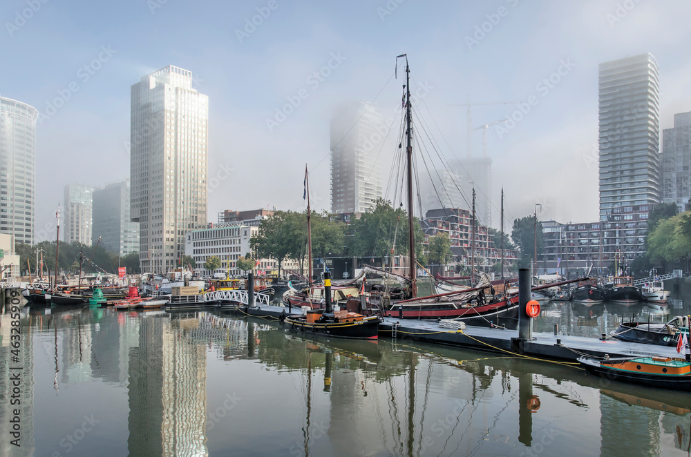 Rotterdam, The Netherlands, October 8, 2021: modern highrise surrounded by patches of mist, reflect in Leuvehaven harbour with its historic vessels