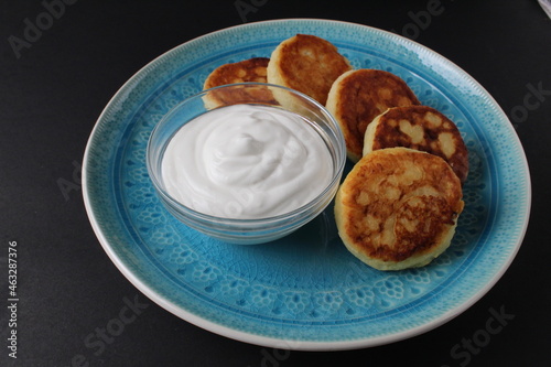 breakfast cheesecakes curd pancakes a few five pieces lie on a large blue dish plate with white sauce sour cream. Traditional dish breakfast Russian Slavic on a black background